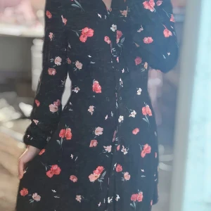 Svart blommig långklänning H&M - Svart långklänning från H&M med rosa och röda blommor. Klänningen har lång ärm, knappar hela vägen framtill och klassisk krage. Skön passform och flowig kjol, perfekt för dig som gillar romantiska vibes och blommigt.