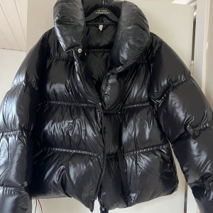 Svart pufferjacka med hög krage - Puffer jacka från Filippa K, modell Gloss Puffer Jacket. Storlek XS. Var inne på kemtvätt förra året men har lite sminkfläckar på insidan av kragen, se bild. Nypris 6700kr.