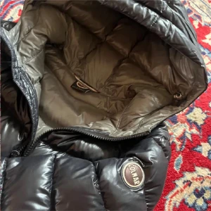 Svart Colmar pufferjacka med huva - Säljer en svart pufferjacka från Colmar med glansig finish och huva. Jackan har ett klassiskt quiltat mönster, dragkedja framtill och Colmar-logga på ärmen. Perfekt för kalla dagar och riktigt snygg till streetwear-stilen.