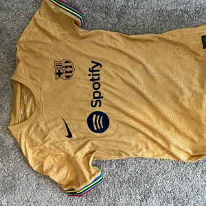 Barcelona guld fotbollströja Nike - Säljer en officiell FC Barcelona fotbollströja från Nike i guld med mörkblå detaljer. Tröjan har korta ärmar med färgglada ränder vid ärmslut, klubbmärke och sponsortryck på bröstet samt UNHCR-tryck på ryggen. Materialet är lätt och andas, perfekt för match eller träning.