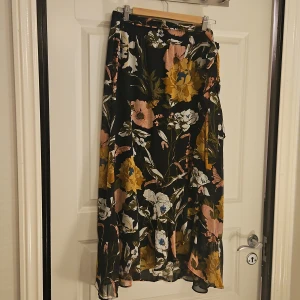 Blommig långkjol från Freequent XL - Säljer en flowy kjol från Freequent i storlek XL. Kjolen har svart botten med stora blommor i gult, rosa och vitt, samt gröna blad. Materialet är lätt och luftigt, perfekt för att skapa en chill vibe. Kjolen har omlott-detalj och volang längs nederkanten.