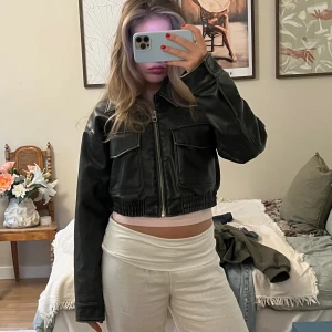 Svart croppad skinnjacka - Säljer en svart croppad skinnjacka med dragkedja framtill och två stora fickor på bröstet. Jackan har bred krage, långa ärmar och elastisk midja för en snygg passform. Perfekt för dig som vill ha en edgy och trendig look.