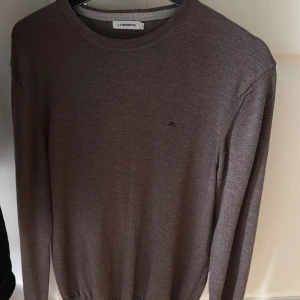 Brun stickad tröja från J.Lindeberg - Snygg brun stickad tröja från J.Lindeberg med rund halsringning och diskret JL-logo på bröstet. Tröjan har lång ärm och en klassisk, stilren look som passar perfekt till jeans eller chinos.
