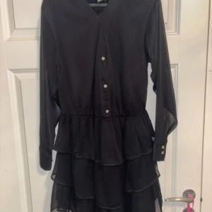 Svart volangklänning med knappar - Svart kortklänning med långa ärmar och flera lager volanger i kjolen. Klänningen har v-ringning och knappar framtill samt transparenta ärmar för en snygg look. Perfekt för dig som gillar stilrena och trendiga plagg.