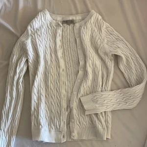 Vit kabelstickad kofta Basic You - Snygg vit kabelstickad kofta från Basic You i ekologisk bomull. Koftan har rund hals, långa ärmar och knappar framtill. Den ribbade kanten vid ärmslut och nederkant ger en clean look. Perfekt för dig som gillar klassisk stil med twist.