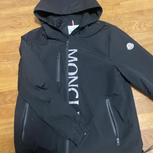 Svart vindjacka från Moncler med huva - Svart vindjacka från Moncler med stor logga framtill, huva och tre dragkedjeförsedda fickor. Jackan har Moncler-märke på ärmen och diskret trikolor-detalj på ryggen. Perfekt för dig som vill ha en clean och stilren look.