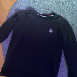 Svart Moncler sweatshirt i storlek S - Svart sweatshirt från Moncler i storlek S med rund hals och långärmad passform. Tröjan har Moncler-logga broderad på bröstet och en stilren design utan mönster eller extra detaljer. Materialet känns mjukt och har en lätt stretch. PS QR KODEN HAR BLIVIT BORT TVÄTTAD 
