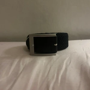 Svart flätat bälte med silverfärgat spänne - Snyggt svart bälte med bred flätad design och ett stilrent, fyrkantigt spänne i silverfärg. Perfekt accessoar för att lyfta din outfit med en modern touch. Bältet har en klassisk look och passar till många olika stilar. 