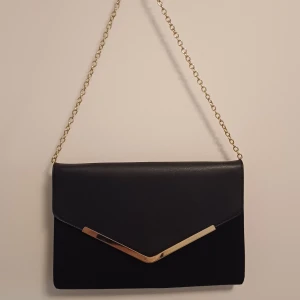 Svart kuvertväska med guldkedja - Stilren svart kuvertväska med guldfärgad kedja och snyggt V-format metallspänne framtill. Väskan har ett innerfack med dragkedja. Perfekt för dig som vill ha en elegant accessoar med plats för det viktigaste.