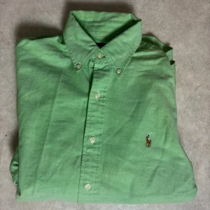 Grön skjorta från Ralph Lauren - Snygg grön skjorta från Ralph Lauren med klassisk button-down krage och broderad logga på bröstet. Skjortan har långa ärmar, vita knappar och är tillverkad i bomull. Perfekt för dig som gillar stilrena och tidlösa plagg.