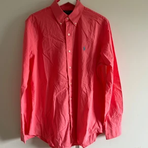 Rosa skjorta från Polo Ralph Lauren - Snygg rosa skjorta från Polo Ralph Lauren med klassisk krage och knäppning framtill. Skjortan har lång ärm och ikonisk blå broderad logga på bröstet. Tillverkad i mjuk bomull och passar perfekt för en stilren look.
