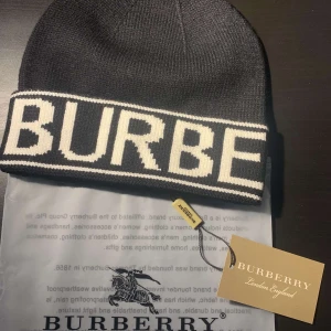 Svart mössa från Burberry med logga - Svart stickad mössa från Burberry med vit, bred logotyp runt hela uppviket. Klassisk och stilren design, perfekt för dig som vill ha en lyxig accessoar med tydlig branding. Materialet är mjukt och stretchigt för skön passform.