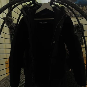 Svart pufferjacka Supply & Demand - Svart lång pufferjacka från Supply & Demand med huva och snygg fuskpäls runt kanten. Jackan har dragkedja, knappar och två stora fickor framtill. Tjockt vadderad för extra värme och cool logga på ärmen. Perfekt för kalla dagar. Pris kan disskuteras och jackan är onesize så det varierar.