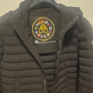 Svart pufferjacka Moose Knuckles L - Svart pufferjacka från Moose Knuckles i storlek L. Jackan har horisontella sömmar, huva med dragsko och flera dragkedjeförsedda fickor. Klassisk logga på ärmen och snyggt broderat innerfoder med färgglada detaljer. Perfekt för kalla dagar. 9/10 skick.