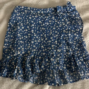 Blå blommig volangkjol med rosett - Supersöt blå kortkjol med vitt blommigt mönster och volangkant🌸 Kjolen har omlottmodell med en rosett i midjan som detalj. Materialet känns lätt och skönt, perfekt för varmare dagar☀️Storlek 140, men sitter perfekt för mig som egentligen har storlek S!❣️ Nästan aldrig använd, och väldigt bra skick!💕