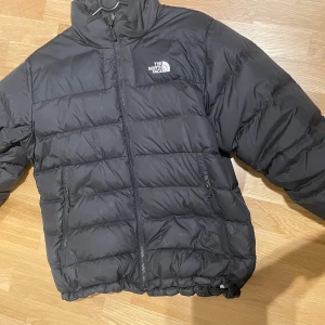Svart pufferjacka från The North Face - Säljer en svart pufferjacka från The North Face i storlek M. Jackan har en klassisk boxig passform, hög krage och två fickor med dragkedja. Perfekt för kalla dagar med sin quiltade design och diskreta logga på bröstet och ryggen.