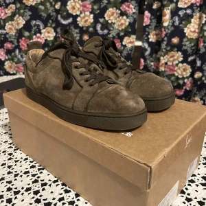 Bruna mockasneakers från Louboutin - Snygga bruna sneakers i mocka från Louboutin med klassisk röd sula. Skorna har rund tå, svarta snören och diskreta sömdetaljer. Perfekta för dig som vill ha en stilren men ändå lyxig look. Kommer med originalkartong.