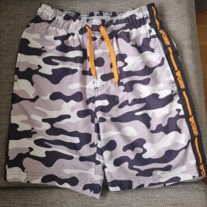 Camo badshorts med snörning från Woxo - Snygga camouflagemönstrade badshorts från Woxo med elastisk midja och orange snörning. Sidorna har svarta band med orange Woxo-logga. Färgerna går i svart, grått och vitt. Det är en fläck på vänster ficka som inte har gått bort i tvätten. Men den är liten och diskret, inget jag tänkt på tidigare. Det är även en reva på badbyxornas bakdel. Tyget är helt, det är bara en liten "skönhetsfläck". Fickorna är även väldigt luddiga men det är ju enkelt åtgärdat (se bilder). Stl 146/152