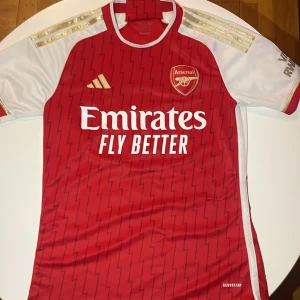 Arsenal hemmatröja Adidas S - Säljer en officiell Arsenal hemmatröja från Adidas i storlek S. Tröjan är röd med vita korta ärmar och har guldiga detaljer på axlarna. Materialet är lätt och andas, perfekt för match eller träning. Zigzag-mönster över hela framsidan och broderat klubbmärke på bröstet.
