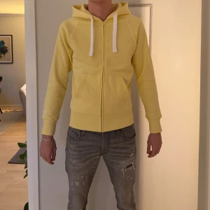 Gul hoodie med dragkedja Russemerch - Fräsch gul hoodie från Russemerch i storlek M. Tröjan har huva med vita snören, praktiska fickor fram och hel dragkedja. Tillverkad i mjuk bomull som är skön mot huden. Perfekt för chill dagar och enkel att matcha med jeans eller joggers.