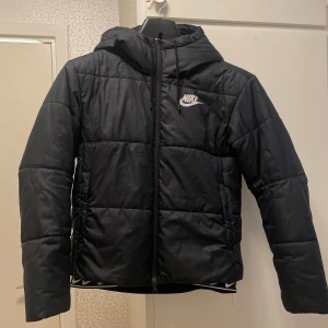 Svart Nike pufferjacka - Snygg svart pufferjacka från Nike med huva och dragkedja framtill. Jackan har quiltad design, lång ärm och klassisk Nike-logga på bröstet.
