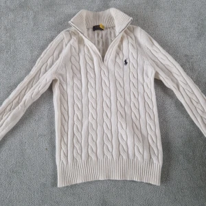 Vit kabelstickad tröja Polo Ralph Lauren - Vit kabelstickad tröja från Polo Ralph Lauren med halv dragkedja och hög krage. Klassisk design med broderad marinblå logga på bröstet. Långärmad och ribbade muddar för en snygg passform. Perfekt för lager-på-lager under kyliga dagar. Storlek S personen på bilden är 181 cm 65 kg. 