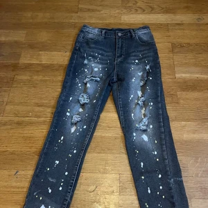 Mörka jeans med färgstänk och hål - Snygga mörkgrå jeans med slitna hål och coola färgstänk i vitt och gult längs benen. Modellen har klassisk femficksdesign och raka ben. Perfekt för dig som gillar en edgy och kreativ streetstil.