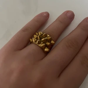 Guldfärgad ring med nitar - Cool och chunky guldfärgad ring med flera konformade nitar på ovansidan. Ringen har en bred design och sticker verkligen ut med sin edgy stil. Perfekt för dig som gillar att addera lite attityd till din look.