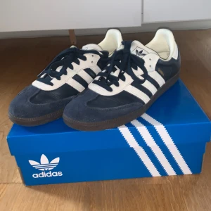 Adidas Samba sneakers marinblå/vit - Adidas Samba sneakers i marinblått och vitt med klassiska tre ränder på sidan, gummisula och snörning. Finns mörkblåa samt beiga snören. Ovandel i skinn och mocka, detaljer i vitt och guldtexten 'SAMBA' på sidan. Perfekta för dig som gillar retrostil och streetwear.