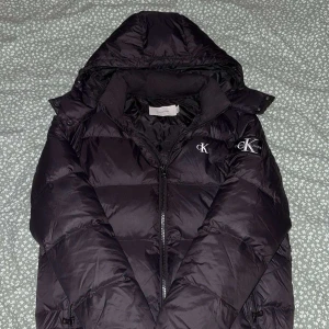 Svart pufferjacka från Calvin Klein - Svart pufferjacka från Calvin Klein Jeans med huva och dragkedja. Jackan har tydliga CK-loggor på bröstet och ärmen. Fylld för extra värme och har en klassisk, lite oversized passform. Perfekt för kalla dagar och riktigt snygg streetstil.