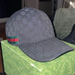  Äkta Svart Gucci keps med GG-mönster - Snygg svart keps från Gucci med klassiskt GG-monogram över hela kepsen och en grön-röd rand på sidan. Kepsen har böjd skärm och är tillverkad i bomullsmaterial. Perfekt för dig som vill ha en exklusiv och trendig look. Och såklart är den äkta och du får med lådan. Storleken är M 58cm