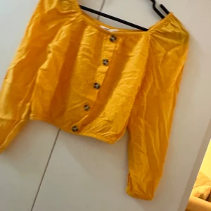 Gul blus med knappar från H&M - Superfin gul blus från H&M med croppad passform och långa ärmar. Blusen har elastisk halsringning som kan bäras offshoulder och snygga knappar framtill. Perfekt för dig som vill sticka ut med färg och stil.