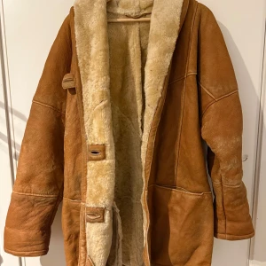 Brun pälsjacka med fårskinnsfoder - Säljer en brun äkta  fårskinns  pälsjacka med mjukt, ljust fårskinnsfoder. Jackan har stora fickor, bred krage och klassiska träknappar. Oversized passform och uppvikta ärmslut för extra chill vibe. Perfekt för kalla dagar när du vill vara både varm och snygg.