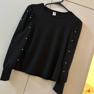 Svart ribbad topp med pärldetaljer SHEIN - Svart långärmad topp från SHEIN med ribbad struktur och pärlor längs ärmarna. Toppen har en rund halsringning och puffiga axlar som ger en trendig look. Perfekt för dig som gillar stilrena detaljer och vill sticka ut lite extra.