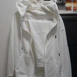 Vit hoodie från Polo Ralph Lauren - Vit hoodie från Polo Ralph Lauren med klassisk svart logobrodyr på bröstet. Tröjan har huva med snörning, dragkedja framtill och stora fickor. Mjuk bomullskvalitet och långärmad passform, perfekt för chill dagar.