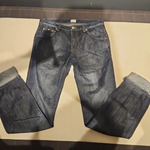 Mörkblå jeans från Armani Jeans - Snygga mörkblå jeans från Armani Jeans med klassisk femficksdesign och guldfärgade nitar. Jeansen har raka ben och tydliga kontrastsömmar. Bakfickan pryds av den ikoniska Armani-loggan i metall. Perfekt för dig som gillar stilrena och exklusiva jeans.
