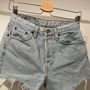Ljusblå jeansshorts från MGB Jeanswear - Säljer ett par ljusblå jeansshorts från MGB Jeanswear med klassisk femficksdesign och råa, fransiga kanter. Modellen är högmidjad och har traditionell knapp och dragkedja framtill. Perfekt för dig som gillar vintagevibbar och relaxed stil.