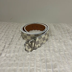 Rutig beige bälte - Snyggt bälte från HM med klassiskt rutigt mönster i beige och grått. Bältet har en stilren spänne i silverfärgad metall och är tillverkat i skinn. Perfekt accessoar för att lyfta din outfit.