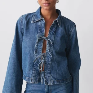  Trendig Blå jeansjacka med snörning - Säljer en blå jeansjacka med snörning framtill och krage. Jackan har en croppad passform och långa ärmar, perfekt för dig som gillar en cool och avslappnad stil. Materialet är klassiskt denim och jackan har en snygg, enkel design. Aldrig använd