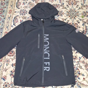 Svart vindjacka från Moncler - Svart vindjacka från Moncler med huva och stora logotypen tryckt vertikalt på framsidan. Jackan har tre dragkedjeförsedda fickor och Moncler-märke på vänster ärm. Snygg och stilren design, perfekt för dig som gillar exklusiva streetwear-vibes.