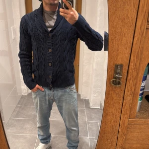 Marinblå kofta från Hampton Republic - Säljer en marinblå kofta från Hampton Republic i storlek S. Koftan har grovstickat flätmönster, ribbade muddar och knappar framtill. Den har en schysst krage och långärmad passform, perfekt för dig som gillar klassisk stil med lite edge.