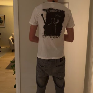 Vit t-shirt The Nights från Avicii - Säljer en vit t-shirt från Avicii med tryck på ryggen som visar en siluett med gitarr och texten 'The Nights'. T-shirten har rund hals och korta ärmar. Materialet är mjuk bomull, perfekt för dig som gillar musik och stilrena prints.