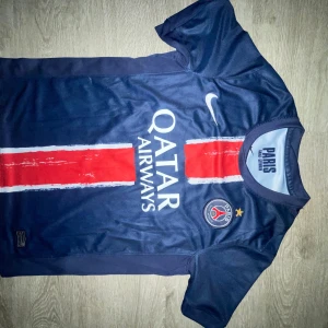PSG matchtröja Nike blå röd - Paris Saint-Germain matchtröja från Nike i blått med röd vertikal rand och vita detaljer. Tryck med Qatar Airways, PSG-emblem och nummer 25 på ryggen. Tillverkad i lätt, ventilerande Dri-FIT-material. Perfekt för dig som älskar fotboll och PSG.
