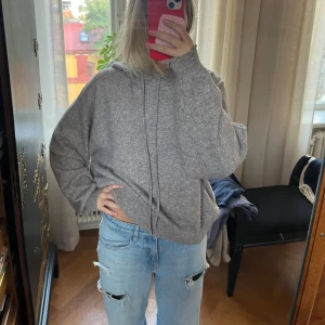 Grå stickad hoodie med huva - Mysig grå stickad hoodie med huva och snörning. Tröjan har en loose passform och långa ärmar, perfekt för chill dagar. Materialet är mjukt och ger en avslappnad vibe. Passar dig som gillar enkel och stilren look.