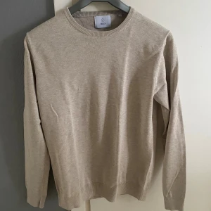 Beige stickad tröja från Bläck - Snygg beige stickad tröja från Bläck i storlek M. Tröjan har rund halsringning och långa ärmar med ribbade muddar vid hals, ärmslut och nederkant. Perfekt för dig som gillar stilrena och enkla plagg med skön passform.