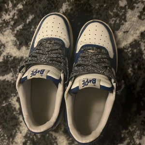 BAPE STA blåvit - Säljer ett par BAPE STA sneakers i blått och vitt med glansig finish och klassisk stjärndetalj på sidan. Skorna har rund tå, platt sula och unika svarta snören med paisley-mönster. Perfekt för dig som vill sticka ut med streetwear och hypebeast-vibe.