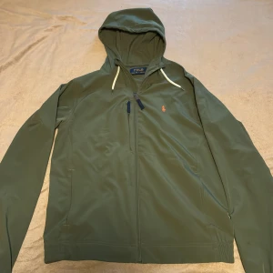 Olivgrön vindjacka från Polo Ralph Lauren - Snygg olivgrön vindjacka från Polo Ralph Lauren med huva och vita dragsnören. Jackan har dragkedja framtill, bröstficka med dragkedja och klassisk röd logga på bröstet. Perfekt för blåsiga dagar och enkel att matcha med det mesta.