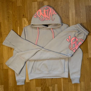 Syna world tracksuit storlek S - Syna World Tracksuit – grå/rosa Snyggt set i grått med rosa detaljer och logga. Hoodie + byxor ingår. Varm och skön för vintern.