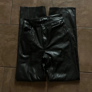 Svarta  byxor i skinnimitation STAY XS - Snygga svarta byxor från STAY i skinnimitation, modell med raka ben och high waist. Byxorna har klassisk femficksdesign, bälteshällor och dragkedja med knapp framtill. Perfekt för dig som gillar en edgy och trendig look. Normala i storleken. 