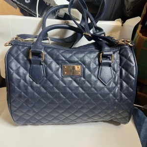 Mörkblå quiltad handväska i skinn - Quiltad mörkblå handväska i skinn med guldfärgade metalldetaljer och dubbla handtag. Väskan har även en avtagbar axelrem och dragkedja upptill. Klassisk och rymlig modell, perfekt för dig som vill ha plats med allt du behöver.